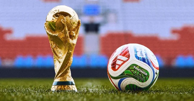 A gold trophy and a white and blue soccer ball are on a vibrant green field, with the trophy slightly to the left and the ball to the right. The background features a blurred stadium with red and blue seats, suggesting a soccer match. The image captures a moment of triumph and celebration.