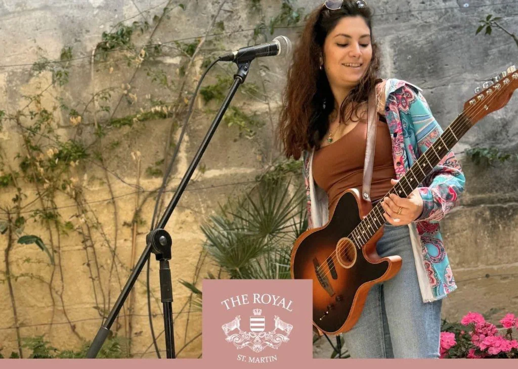 A woman in a brown jacket and blue jeans plays a brown guitar on stage, with a microphone to her left and a "The Blue Guitar" banner in the background. The scene is set against a stone wall with plants, creating a relaxed and intimate atmosphere.