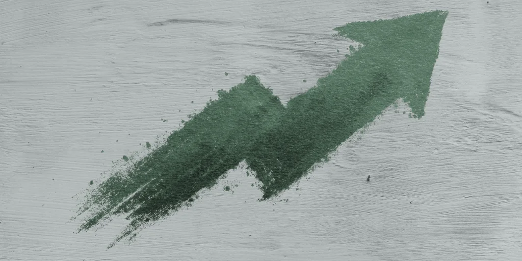 A green, textured arrow with a jagged tip is centrally positioned on a gray wooden surface, creating a dynamic and visually striking composition. The arrow's position and the wooden surface's texture contrast, while the green color adds a pop of color. The image conveys a sense of movement and direction, possibly related to a new product launch or a business concept.