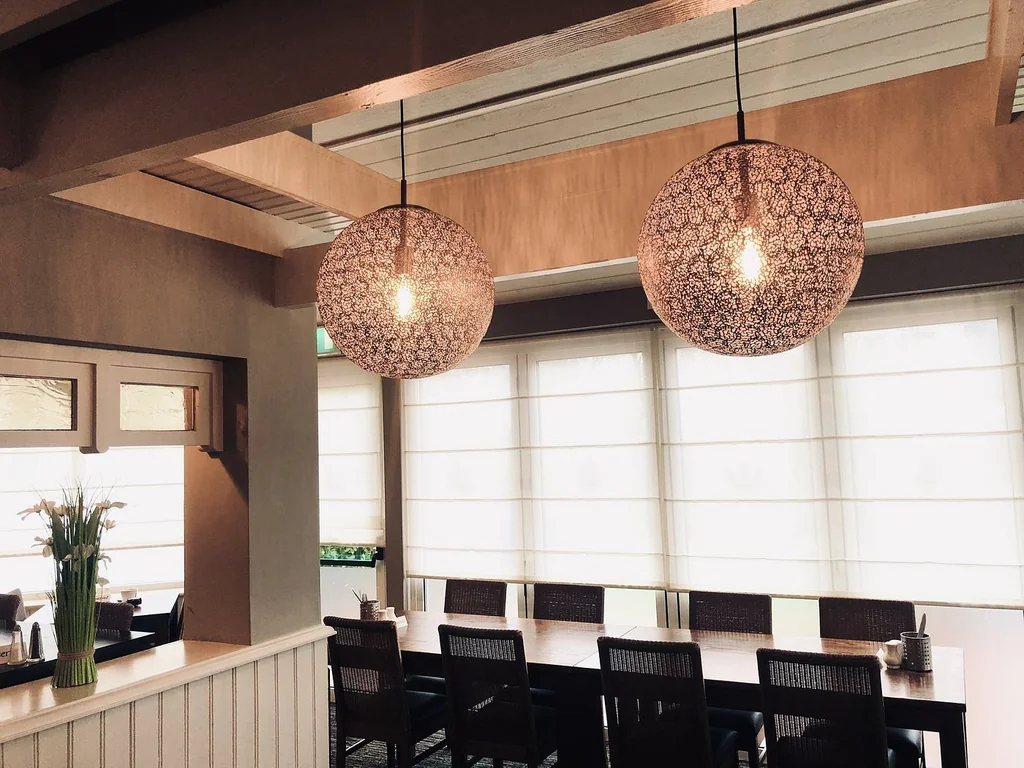 The image features a modern, minimalist dining room with a wooden ceiling, white walls, and a large window with white curtains. Two hanging light fixtures with intricate designs hang from the ceiling, casting a warm glow. The room has a minimalist aesthetic with a wooden counter, black chairs, and a vase of flowers. The arrangement of objects creates a harmonious balance, making the space inviting and comfortable.