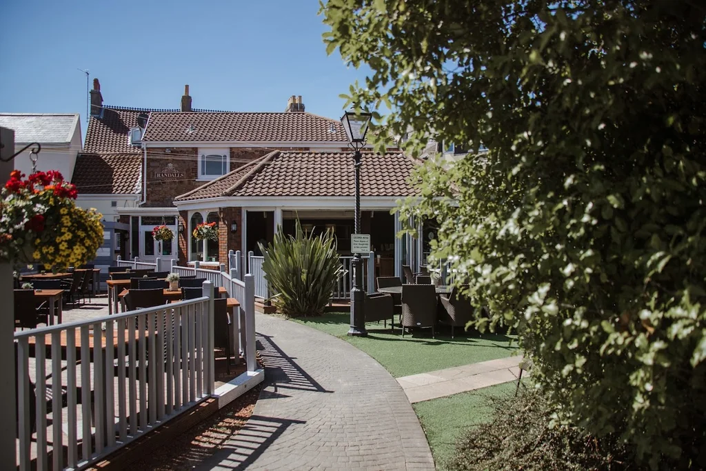 A serene residential area features a white fence, a white picket fence, and a white porch with a wooden table and chairs. A red-roofed building with a white roof and a red roofed shed are in the background. The scene is set against a clear blue sky, with lush green trees and bushes in the foreground. The image is taken from a low angle, emphasizing the building and the surrounding greenery. The landmark identifier "sa_15007" does not provide additional information about the location.