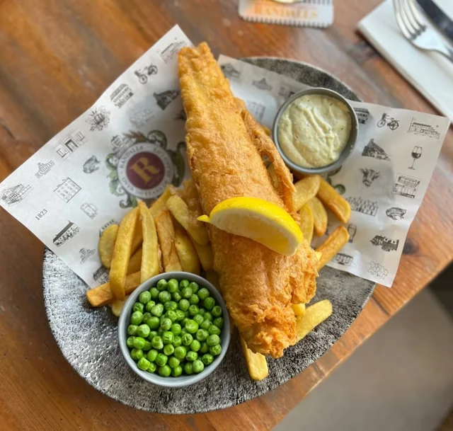 A golden brown fish fillet is the centerpiece of a meal on a black and white plate with a white napkin. The plate is on a wooden table with a white napkin and a fork nearby. Two small bowls of green peas and fries accompany the fish, and a slice of lemon adds a touch of freshness. The scene is visually appealing and inviting.
