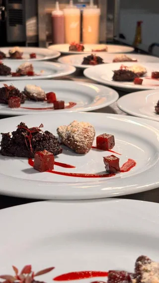 A table displays 12 white plates with various desserts, arranged in two rows. The desserts include chocolate cake, strawberry sauce, and red berry. Red sauce drapes over the desserts, and the desserts are arranged meticulously. The background features a kitchen counter with condiments and a coffee machine, creating a professional atmosphere.