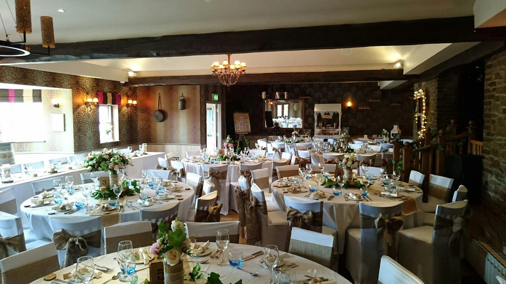 A grand banquet hall is depicted, with tables draped in white tablecloths and adorned with green and white floral arrangements. The room features rustic brick walls, a chandelier, and a fireplace, creating a warm and inviting atmosphere. The arrangement of tables and chairs suggests a formal event, possibly a wedding or reception.