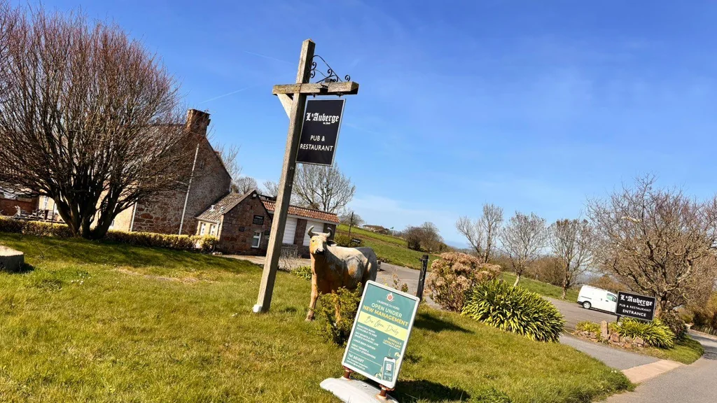 The image features the historic Blueston Inn in the countryside, with a wooden signpost displaying the name and location. The signpost is on a grassy area, with a cow statue nearby. The scene is set against a clear blue sky, and the Blueston Inn is surrounded by trees and buildings. The image captures the peaceful atmosphere of the location.