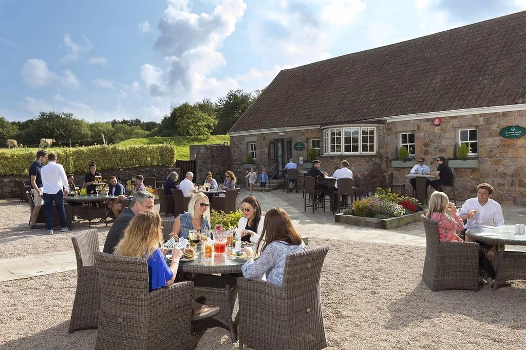 A lively outdoor gathering takes place at a stone pub or restaurant, surrounded by a gravel patio and lush greenery. The patio is filled with people enjoying meals and drinks, while the pub or restaurant has a rustic charm with stone walls and a thatched roof. The scene is set against a backdrop of a clear blue sky and lush greenery, creating a warm and inviting atmosphere.