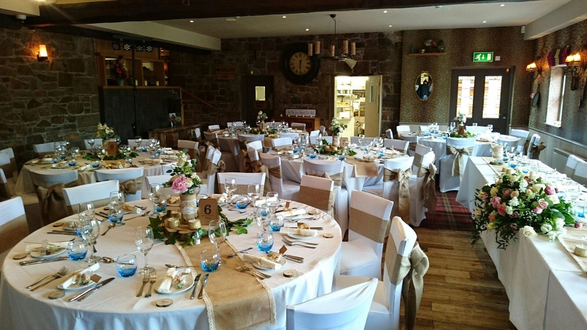 A beautifully set banquet hall features round tables with white tablecloths, blue and white flowers, and silverware. The tables are surrounded by white chairs, and the room has rustic charm with stone walls, a fireplace, and a clock. The space is well-lit and organized, ready for a special occasion.