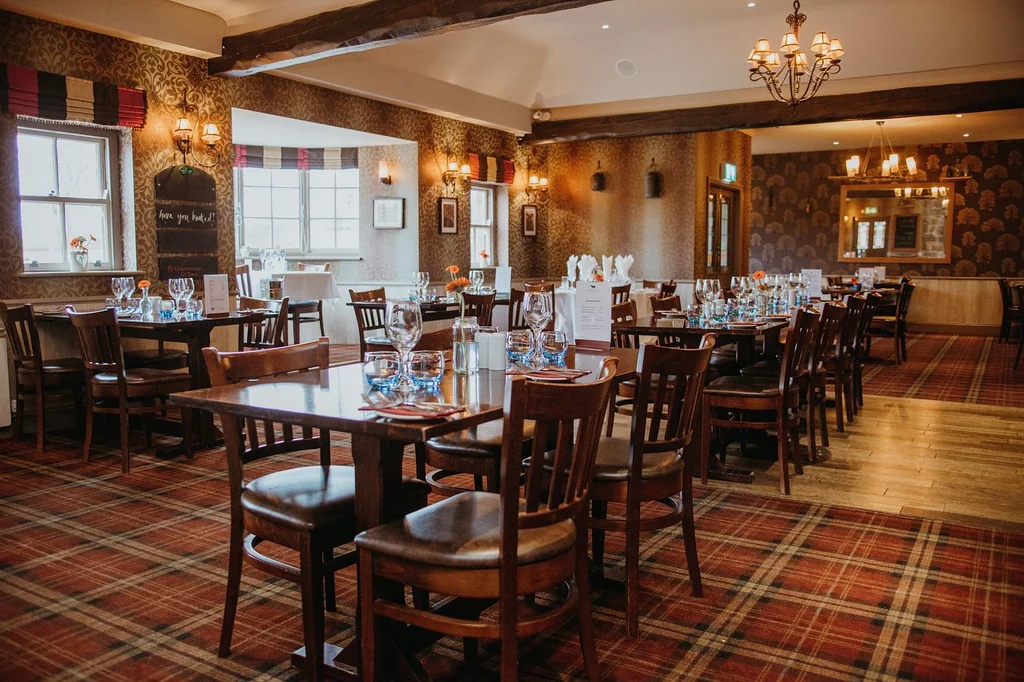 The image shows a cozy, well-lit restaurant with wooden floors and walls, featuring a red and black checkered pattern. Tables are set with white tablecloths and silverware, and chandeliers hang from the ceiling. The walls are adorned with framed pictures and a sign that reads "The Bar". The room is inviting and ready for guests.