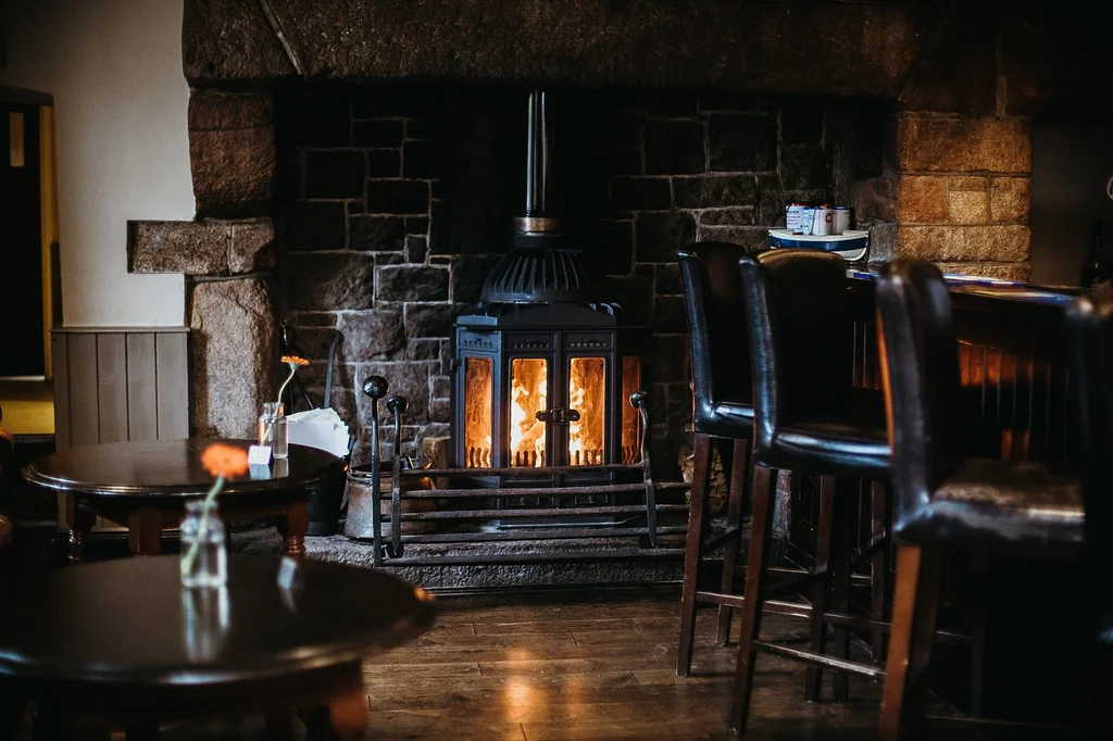 A cozy bar area features a dark stone fireplace with a lit fire, casting a warm glow. A wooden table with a vase of flowers and a wooden stool are in front of the fireplace. Bar stools are scattered around, and a wooden bar is on the left side. The room has a warm, inviting atmosphere with a wooden floor and a stone wall.