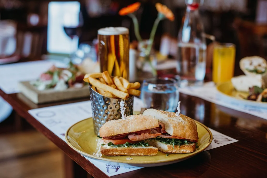 A wooden table features a golden plate with a sandwich, fries, and a glass of water. A white napkin and a yellow cup with a straw are also present. A vase with orange flowers adds color. The scene is inviting and well-prepared for a meal.