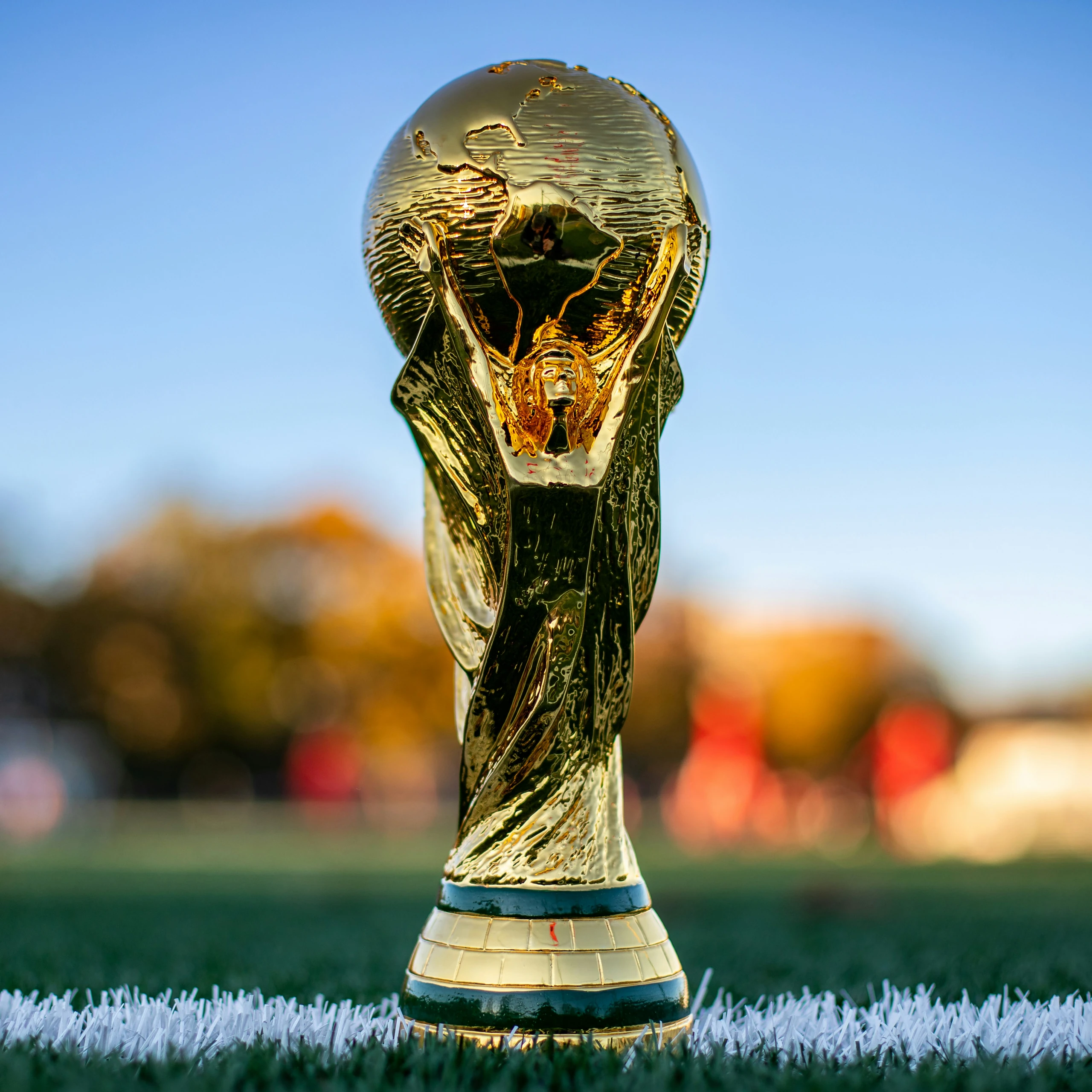 A golden trophy with a human head and wings, adorned with a gold crown, stands on a white soccer field, surrounded by a blurred background of trees and a clear blue sky. The trophy is the central focus, with the field and sky providing context.