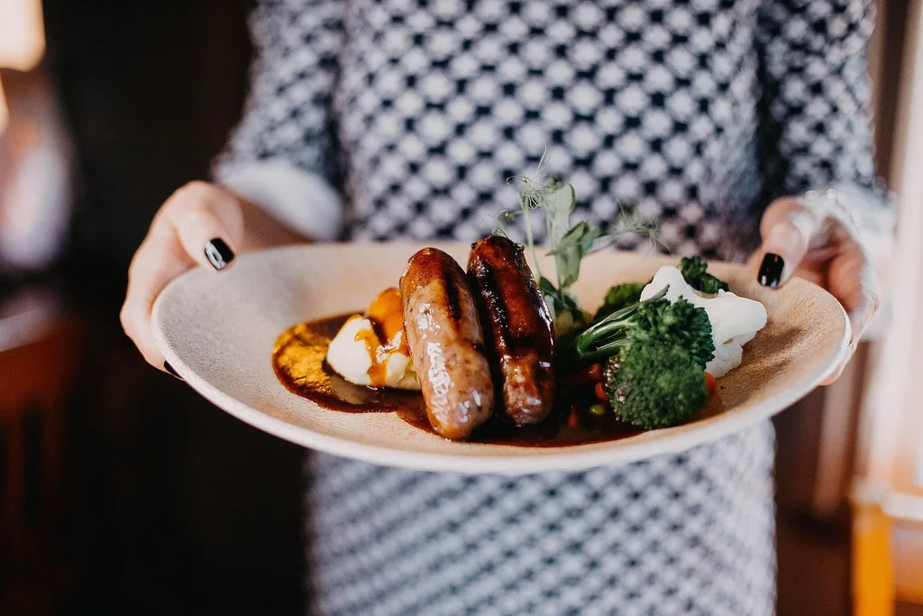 A person in a black and white checkered dress holds a white plate with a variety of food, including sausages, mashed potatoes, and broccoli, in a restaurant or home setting. The plate is on a wooden table, and the blurred background suggests a restaurant or home setting. The person's hands are adorned with black nail polish, and the plate is the main focus.