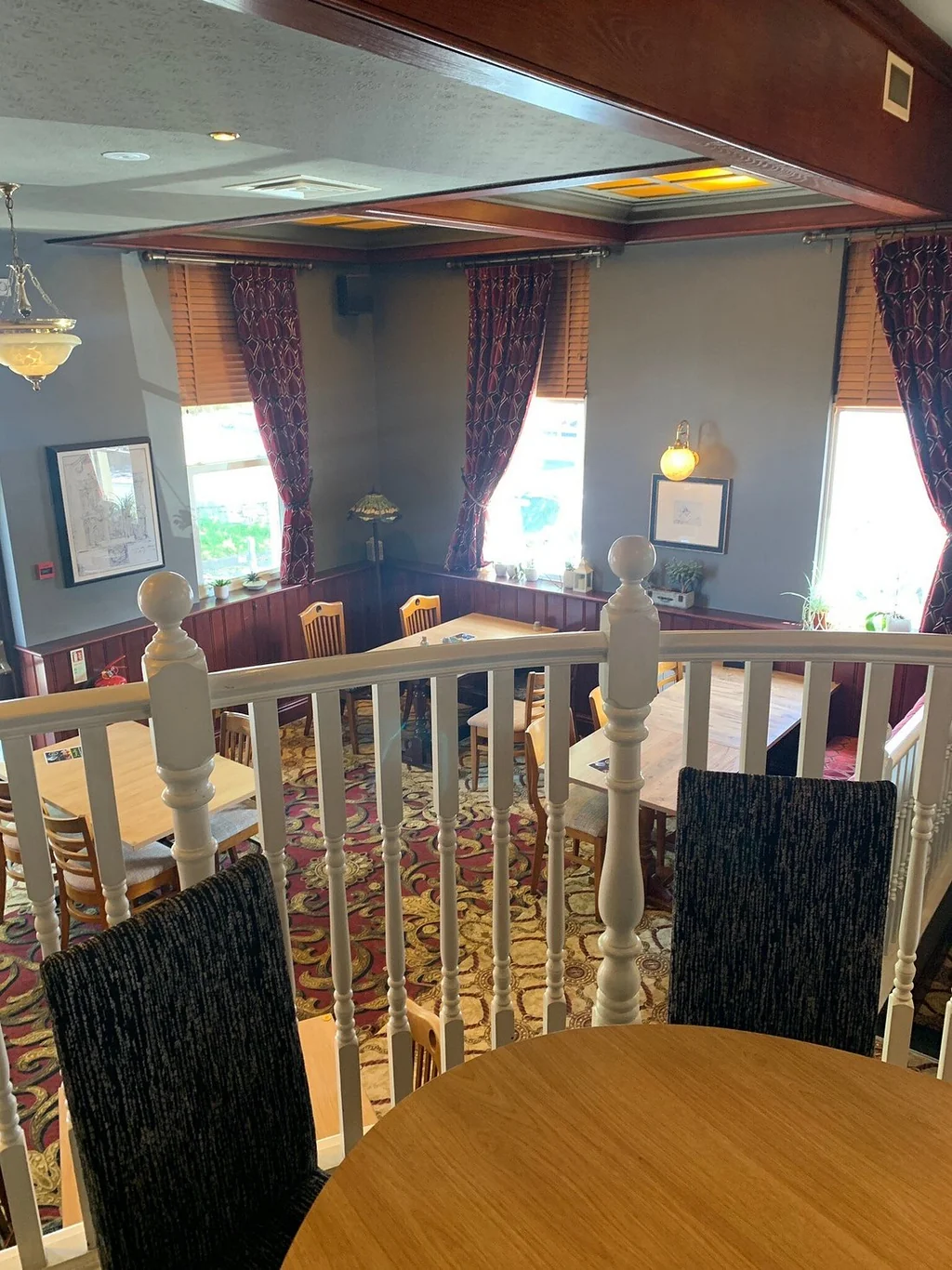 The image shows a cosy dining area with a wooden table and chairs, a white railing, and a wooden floor. The room has a warm, inviting atmosphere with a chandelier and curtains. The perspective is from the balcony, offering a bird's eye view of the dining area. The room is well-lit and has a cosy ambiance.
