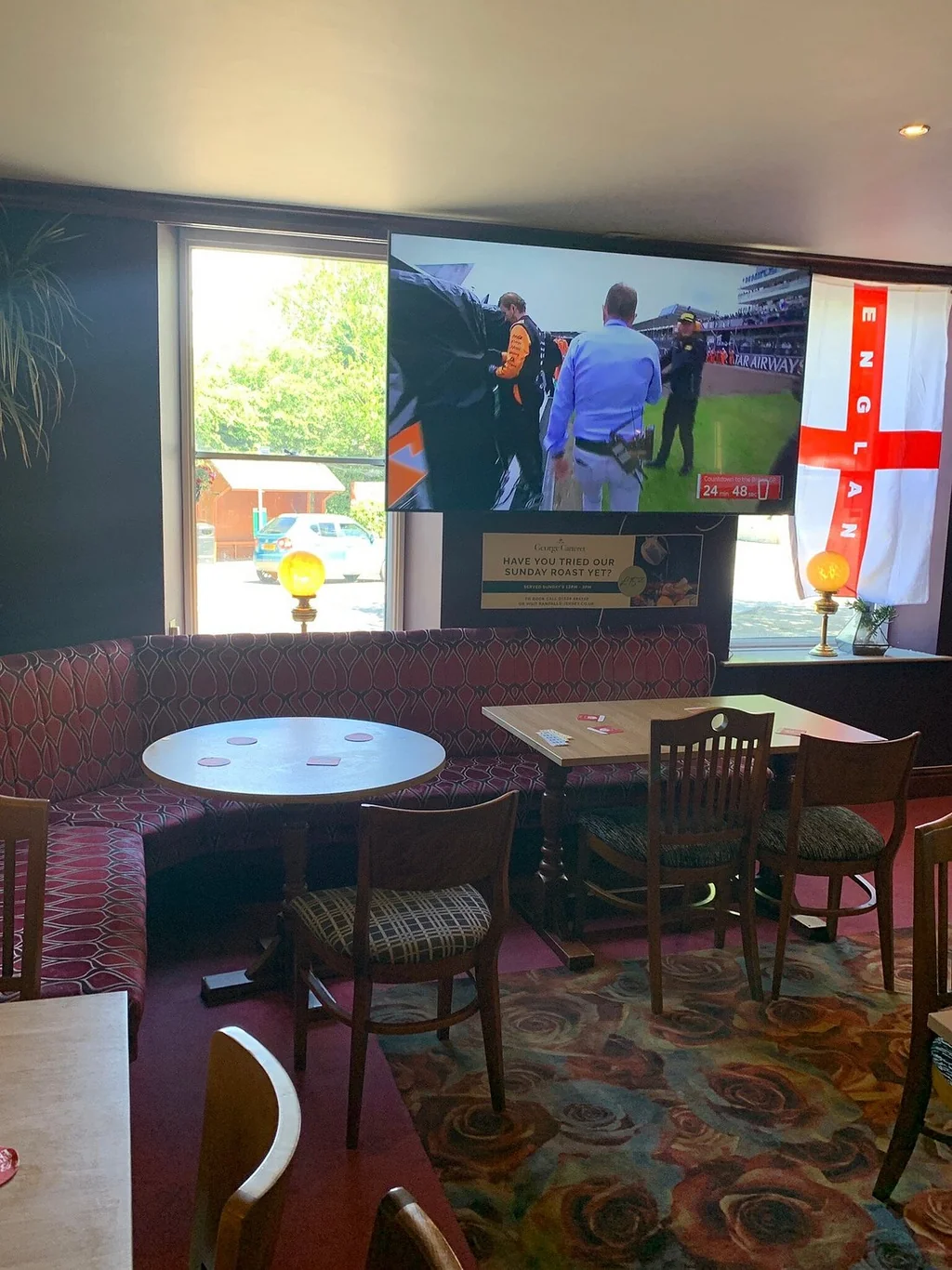 The image shows a cosy, modern restaurant with a red and black patterned carpet, a wooden table with a white tablecloth, and a red sofa with a blue and white striped cushion. The walls are adorned with posters of a soccer game and a football match, and a large window offers a view of a street. The restaurant is well-lit and inviting, with a warm and inviting atmosphere.