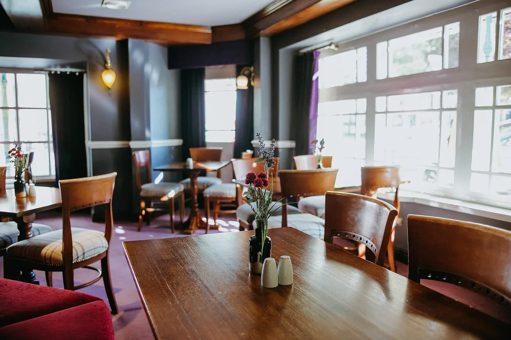 The image shows a cosy restaurant with warm wooden furniture, a central wooden table with a vase of flowers, and a red chair. The walls are painted purple, and the floor is carpeted in dark purple. Large windows with white curtains allow natural light to enter, and a hanging light fixture adds to the ambiance. The restaurant is well-organized and inviting, with a harmonious color scheme and attention to detail.
