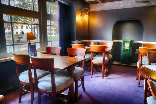 The image shows a cosy, well-lit room with a wooden table, six chairs, and a blue vase with a white flower. The room has a purple carpet, a large window with blue curtains, and a green plant. The walls are painted in a soothing shade of blue, and a chandelier hangs from the ceiling. The room is inviting and cosy, perfect for relaxation or socializing.