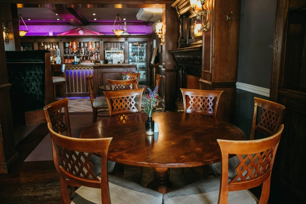 A cosy bar or restaurant features a round wooden table with six chairs, a vase of flowers, and a mirror. The dark wooden walls and ceiling create a warm atmosphere, with a fireplace and chandeliers adding to the ambiance. The scene is inviting and well-organized, with a focus on the table and chairs.