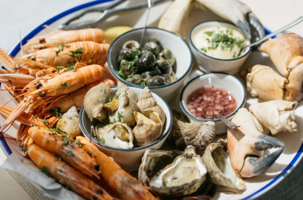 A white plate with a blue rim holds a variety of seafood dishes, including orange prawns, clams, mussels, and oysters. The plate is on a white tablecloth, with a lemon wedge and a small bowl of sauce nearby. The arrangement is visually appealing and suggests a carefully prepared meal.