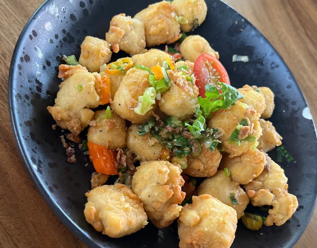 A black plate with golden brown fried chicken pieces, garnished with parsley and cherry tomatoes, sits on a wooden table. The plate is centrally positioned, with the chicken pieces slightly overlapping the plate. The background is blurred, emphasizing the plate and its contents.