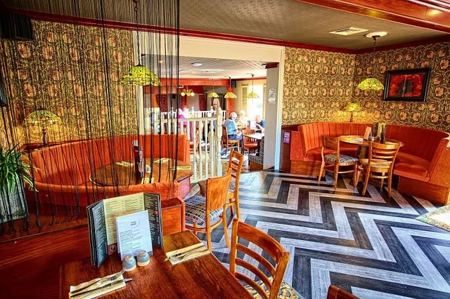 A modern, elegant restaurant with a unique design features a wooden dining table with a wooden chair, a wooden bench, and a wooden table with a wooden chair. The walls are adorned with floral wallpaper, and the ceiling has hanging lights. The floor is covered in black and white checkered tiles. A man is seated at a wooden table, and a woman is seated on a red couch. The restaurant has a cosy atmosphere with a wooden table, wooden chairs, and a wooden bench.