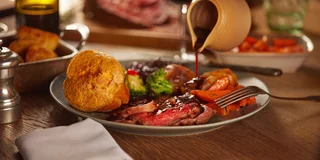 A wooden table features a large, dark brown steak with a red sauce, surrounded by a variety of food items, including a golden-brown roll, a white bowl, a white plate, a silver fork, a glass of wine, and a white napkin. The scene is set against a blurred background, emphasizing the meal's rich colors and textures.