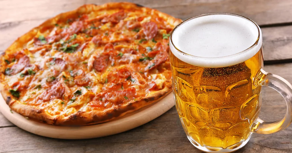 A golden-brown pizza with vibrant toppings sits on a wooden board, accompanied by a tall, frothy beer mug filled with golden beer. The scene is set on a rustic wooden table, creating a cozy atmosphere.