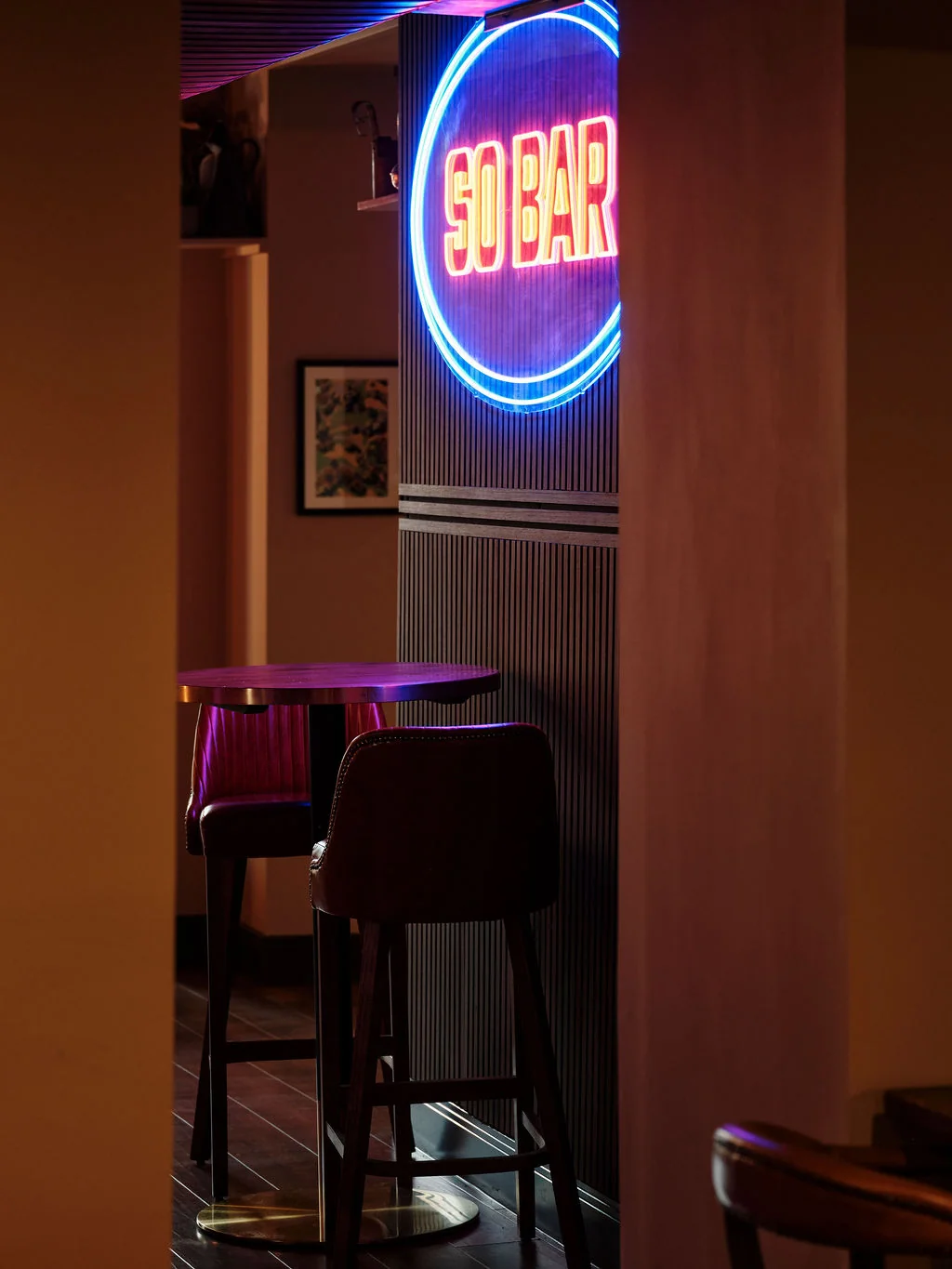 A dimly lit, cosy bar scene features a neon "SAND" sign on a white wall, with two wooden stools and a wooden bar in the foreground. The floor is dark wood, and a painting hangs on the wall. The perspective is from the entrance, inviting patrons to join.