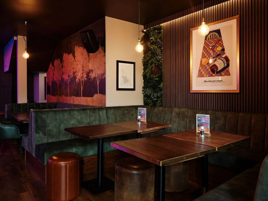 A modern restaurant interior features a wooden floor, green booths, and a wooden bench. The walls are adorned with colorful posters and a large poster with a tree design. The ceiling has hanging lights and a black speaker. The atmosphere is cosy and inviting, with a warm ambiance.