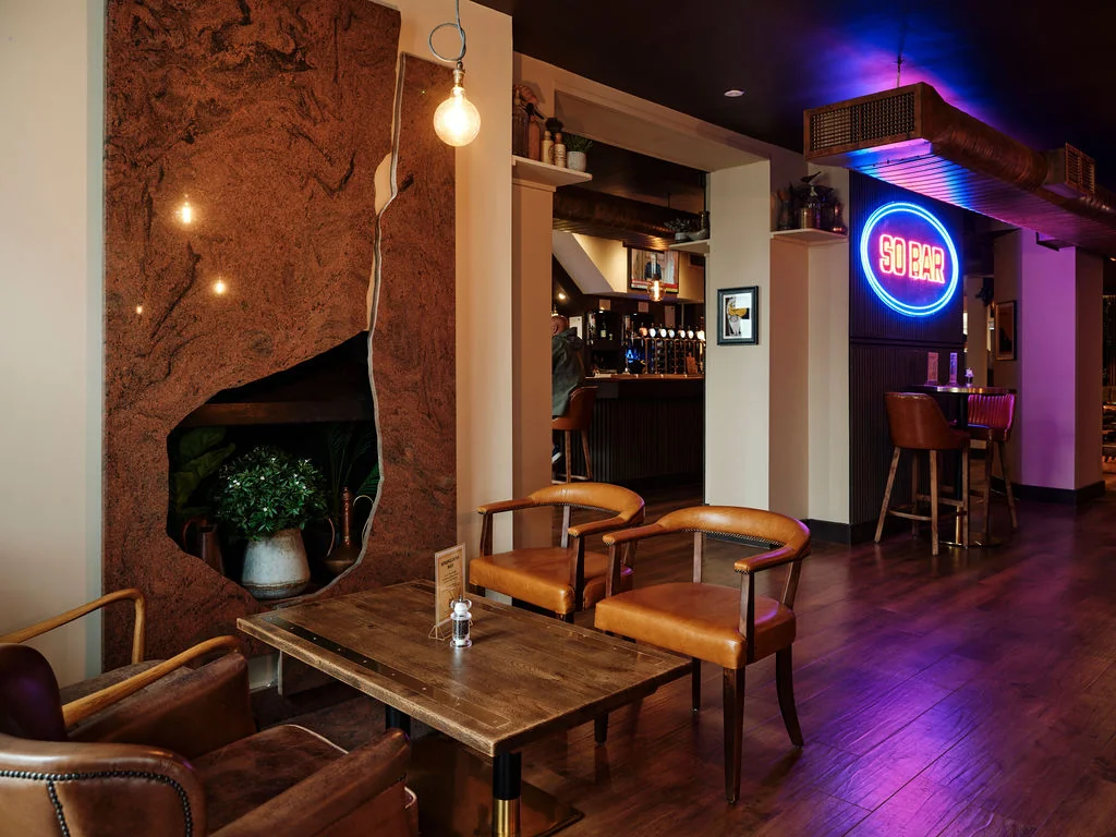 A cosy bar or restaurant features a rustic wooden wall with a large hole, a wooden table with a vase of flowers, and a wooden chair. The bar has a neon sign and a potted plant. The floor is dark wood, and the ceiling has hanging lights. The atmosphere is warm and inviting, with a welcoming ambiance.