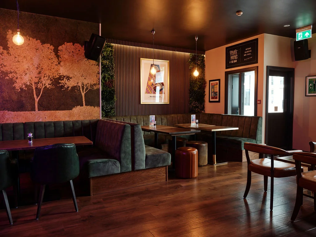 A cosy, rustic restaurant with dark wood floors and green velvet booths is illuminated by hanging lights. A large mural of a tree and a menu sign are on the walls, while a wooden table with a candle and a brown trash can are in the center. A door with a window and a green sign are on the right side, and a hanging light fixture is on the left. The atmosphere is warm and inviting, perfect for a cosy evening.