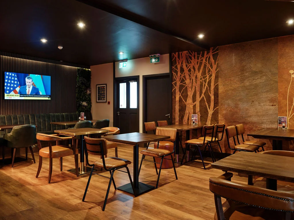 A modern, cosy bar with dark wood floors and a wooden wall features a large flat-screen TV displaying a woman in a blue shirt. The bar has six tables, two green couches, and a wooden bar. A wooden tree mural adds a touch of nature. The bar is well-lit, creating a warm and inviting atmosphere.