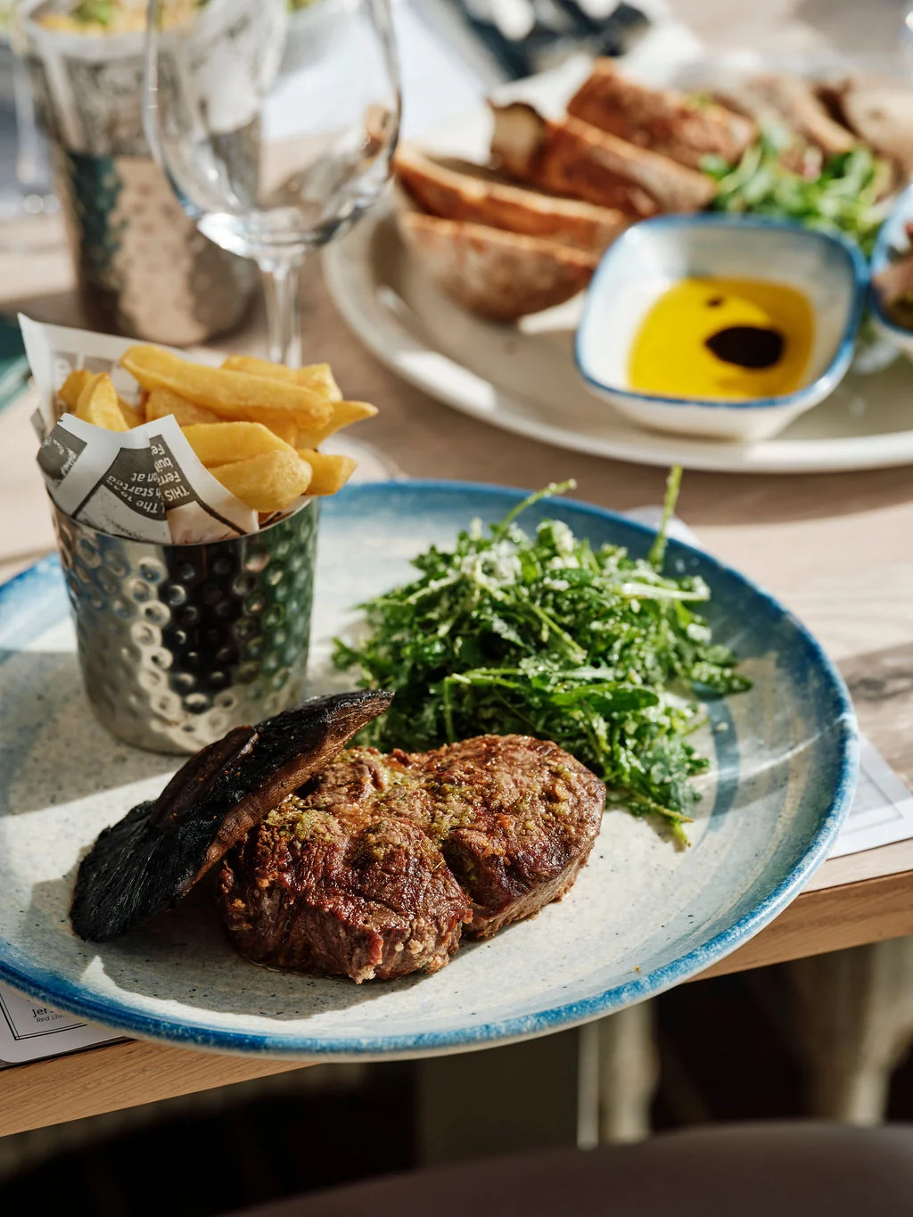 A dining table features a steak with a dark brown crust and green sauce, accompanied by a side of fries and a glass of wine. A white plate with a blue rim holds the steak, while a silver cup with a yellow sauce and a white napkin are nearby. The scene is set against a blurred background, emphasizing the meal's presentation.
