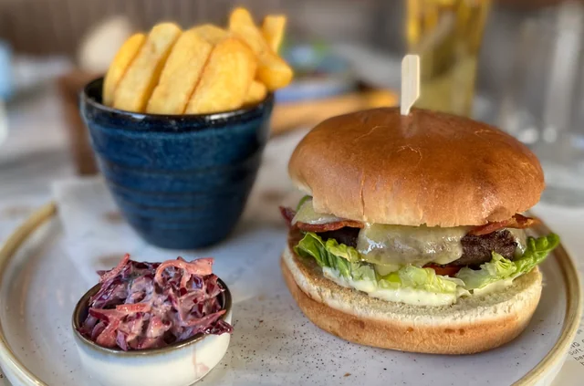 A delectable burger with a golden bun, lettuce, and meat is the centerpiece of the image, held together by a toothpick. Accompanying the burger are golden fries and coleslaw, both served in a small bowl. The meal is set on a white plate, with a blue cup and glass of water in the background. The arrangement suggests a casual dining experience, with the burger as the star.