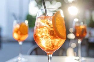 Two glasses filled with orange juice and ice cubes are on a white table, with one straw in each. The background is blurred, suggesting an outdoor setting. The image is taken from a low angle, emphasizing the glasses and their contents. The scene is serene and inviting, with the sun casting a warm glow.