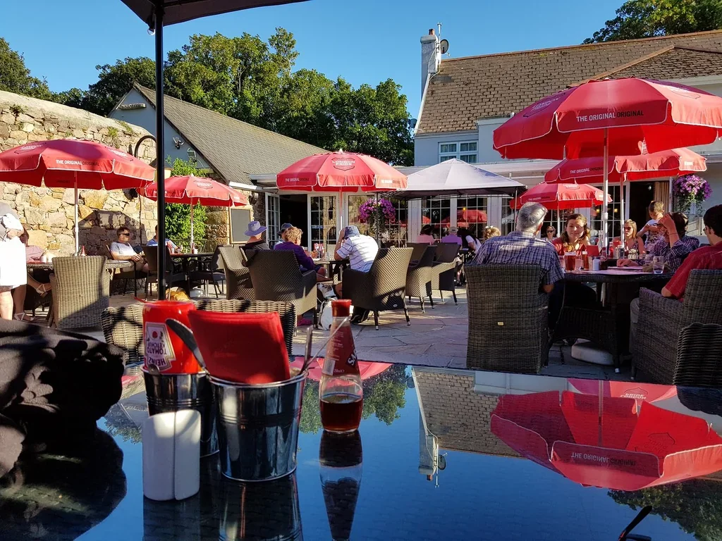 The image shows a lively outdoor café with red umbrellas, wicker chairs, and tables reflecting the surroundings. People are seated and enjoying the ambiance, with some standing. The café is surrounded by a stone wall and a white building with a red roof. The scene is set against a clear blue sky, creating a vibrant and inviting atmosphere.