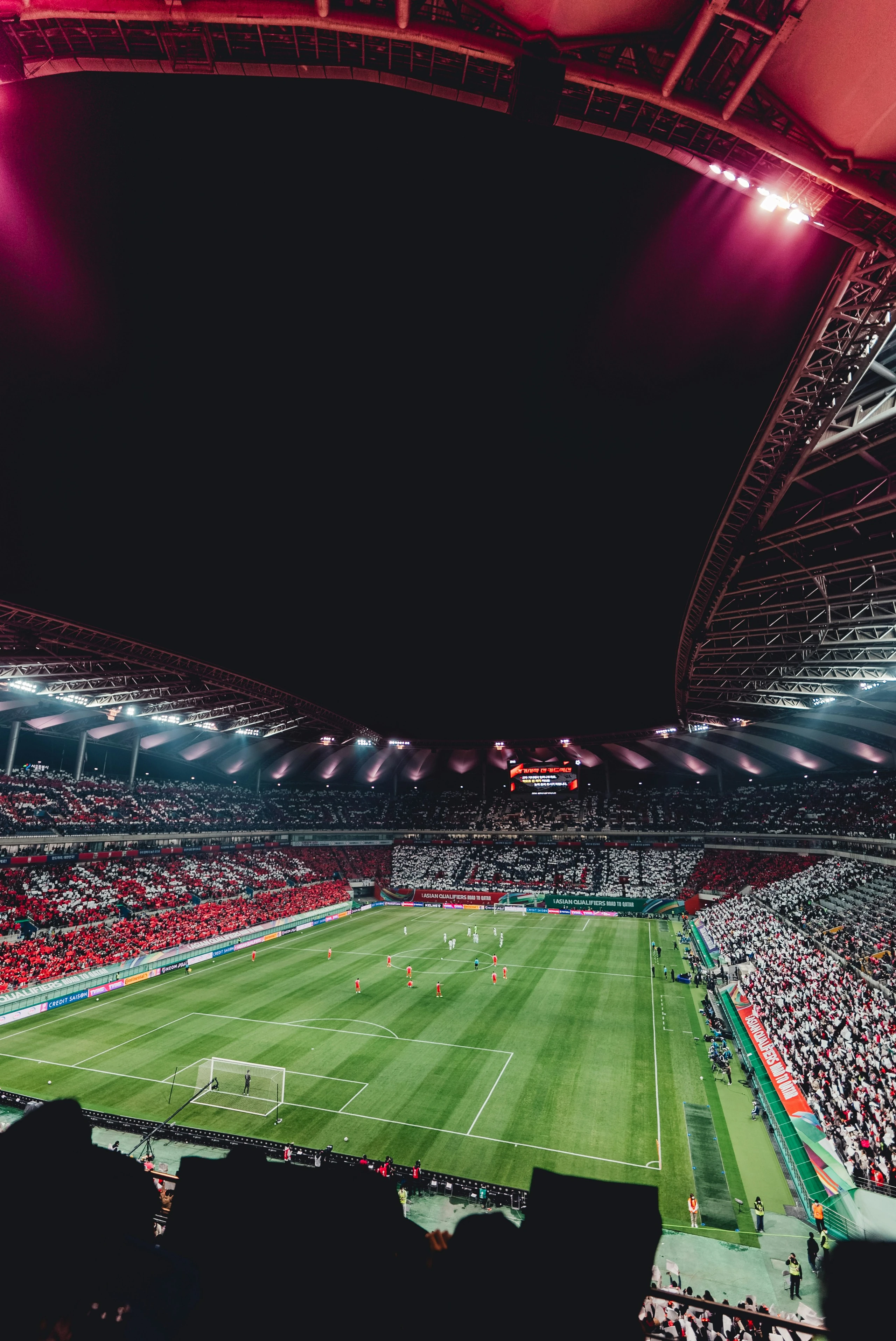 A nighttime soccer match is captured in a large stadium, with a vibrant green field and white boundary lines. Players are scattered across the field, and the stands are filled with excited spectators. The stadium is illuminated by bright lights, and a large scoreboard is visible in the background. The image is taken from the stands, offering a comprehensive view of the match.