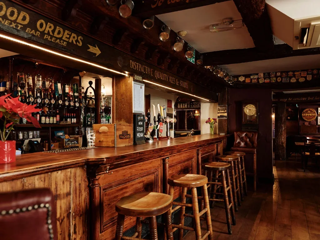 The image shows a cozy, rustic pub with a wooden bar, three stools, and a potted plant. The bar is adorned with bottles and glasses, and a sign above reads "Ollie's Bar". The background features a wall with a clock and a poster, and a ceiling fan. The scene is warm and inviting, with a sense of history and comfort.