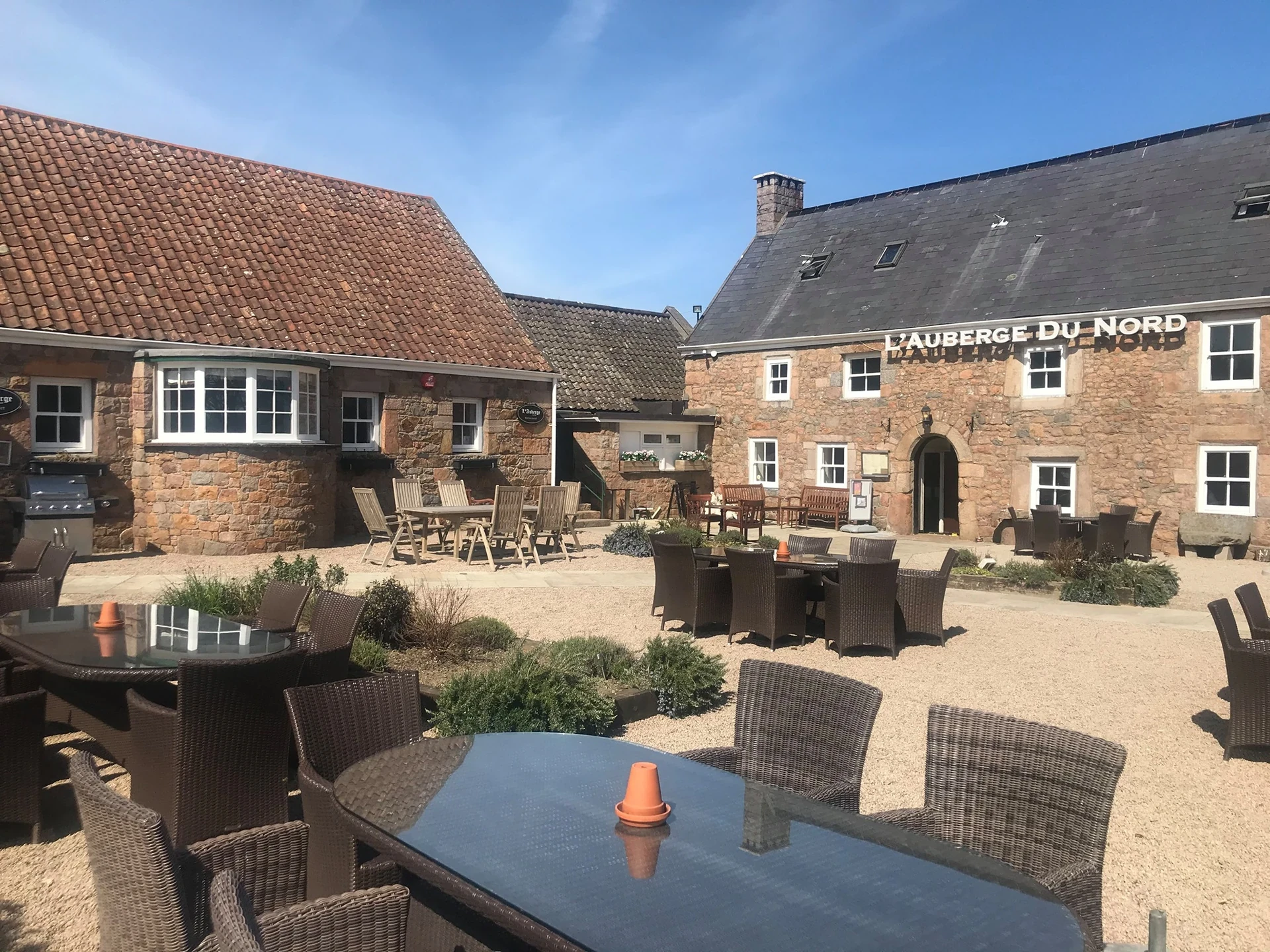 The image shows the historic "Huis de Brouwer" pub in the Netherlands, with a brick building and a red tiled roof. The pub is surrounded by a gravel courtyard, with tables and chairs arranged for patrons. The clear blue sky and greenery create a serene atmosphere, making it a popular spot for locals and tourists alike.