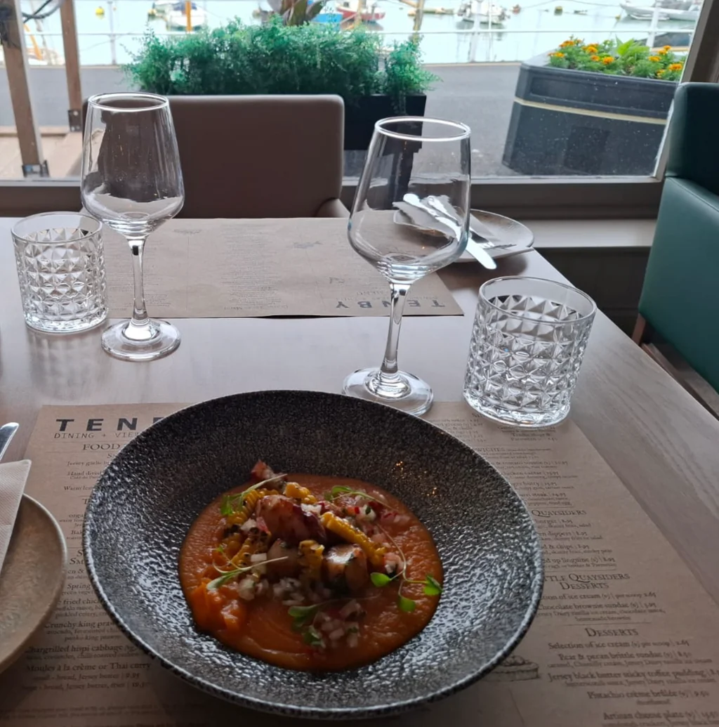 A serene dining scene features a dark gray bowl with a colorful topping on a white tablecloth, accompanied by two empty glasses and a menu. A green plant adds a touch of nature. The view outside the window reveals a harbor with boats, creating a tranquil atmosphere.