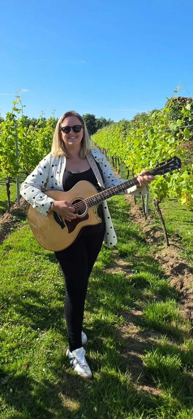 A woman stands in a vineyard, holding an acoustic guitar and posing for a photo. She wears sunglasses and a white and black jacket, with her hair styled in loose waves. The vineyard is lush with vines and trees, and the sky is clear blue. The image captures a serene moment of relaxation and appreciation of nature.